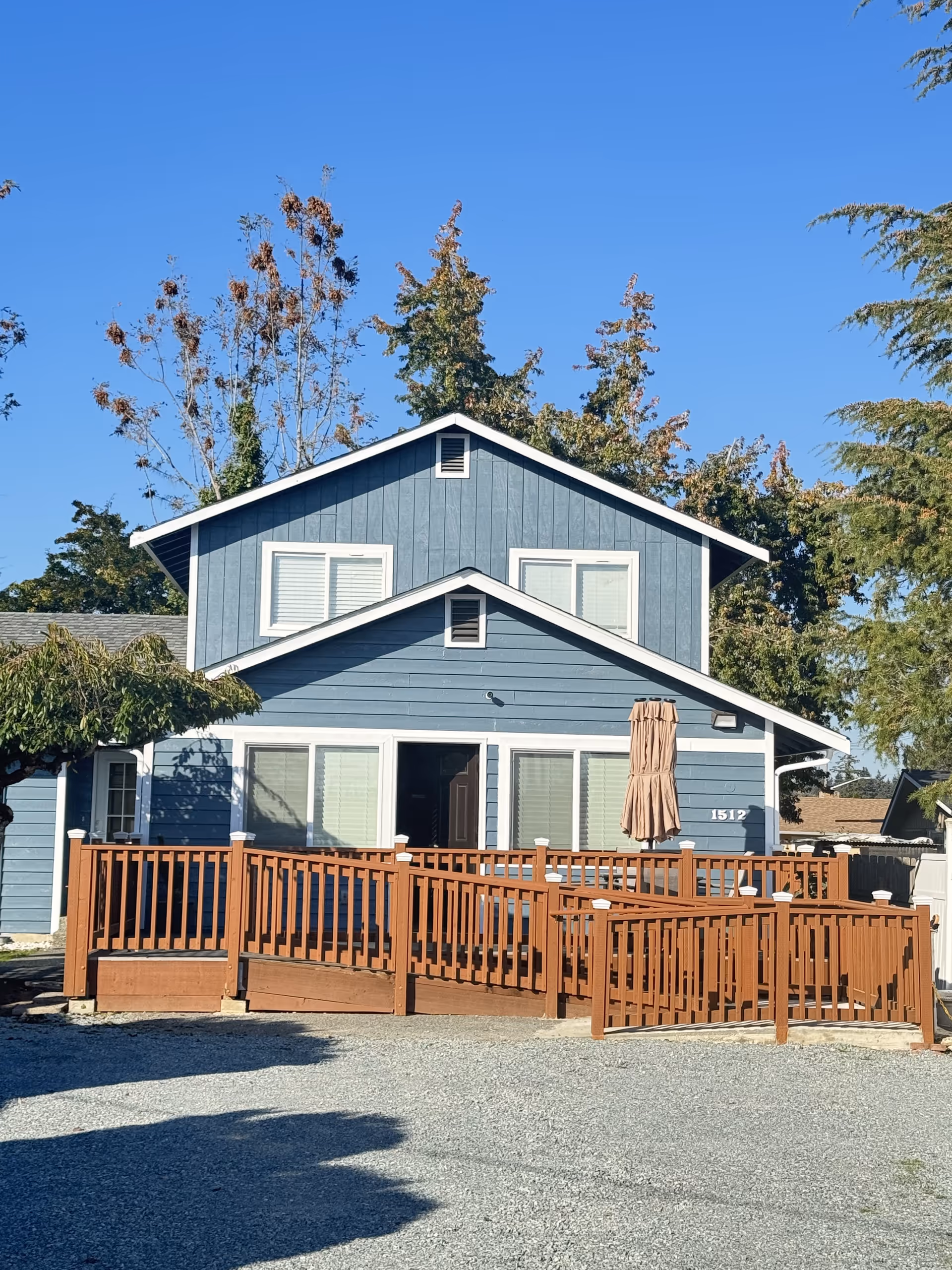 Blue two-story home with a wooden deck, railing, and a closed umbrella.