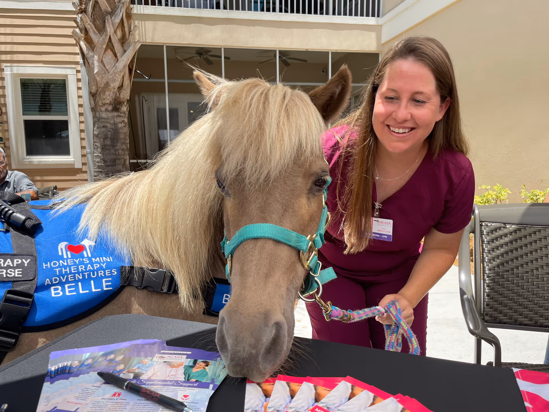 Caregiver in burgundy scrubs smiles near a therapy pony outdoors at ACASA SeniorCare