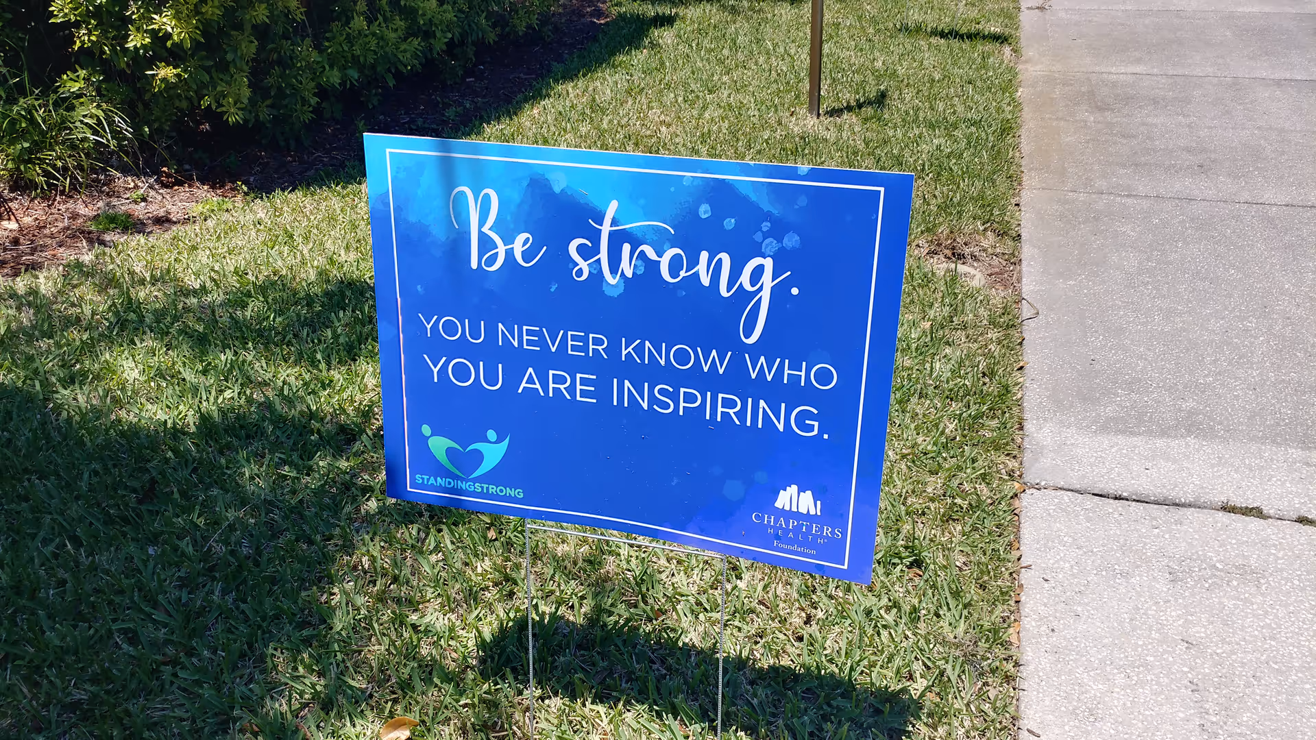 Blue inspirational sign on a grassy lawn beside a sidewalk.