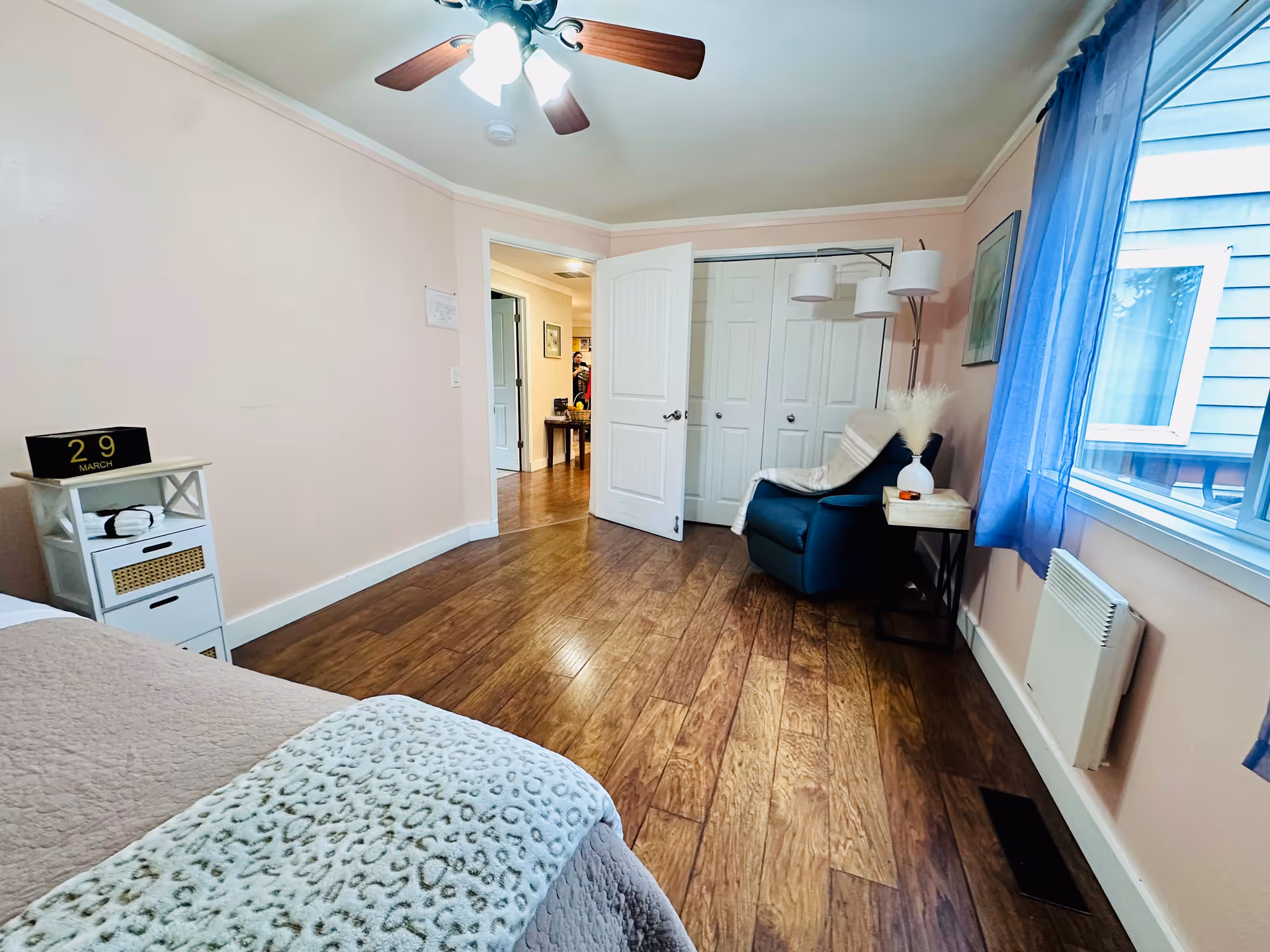 A pink-walled bedroom with a bed, chair, nightstand, blue curtains, and doorway.