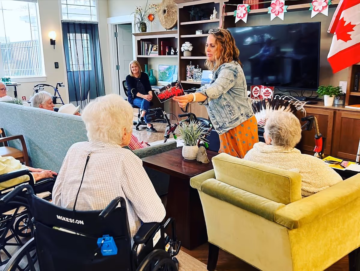 A group of elderly residents seated in a living room area of a senior living facility, attentively listening to a woman standing and showing them a picture. The room has comfortable seating, a large TV, shelves with decorations, and a Canadian flag displayed on the wall.