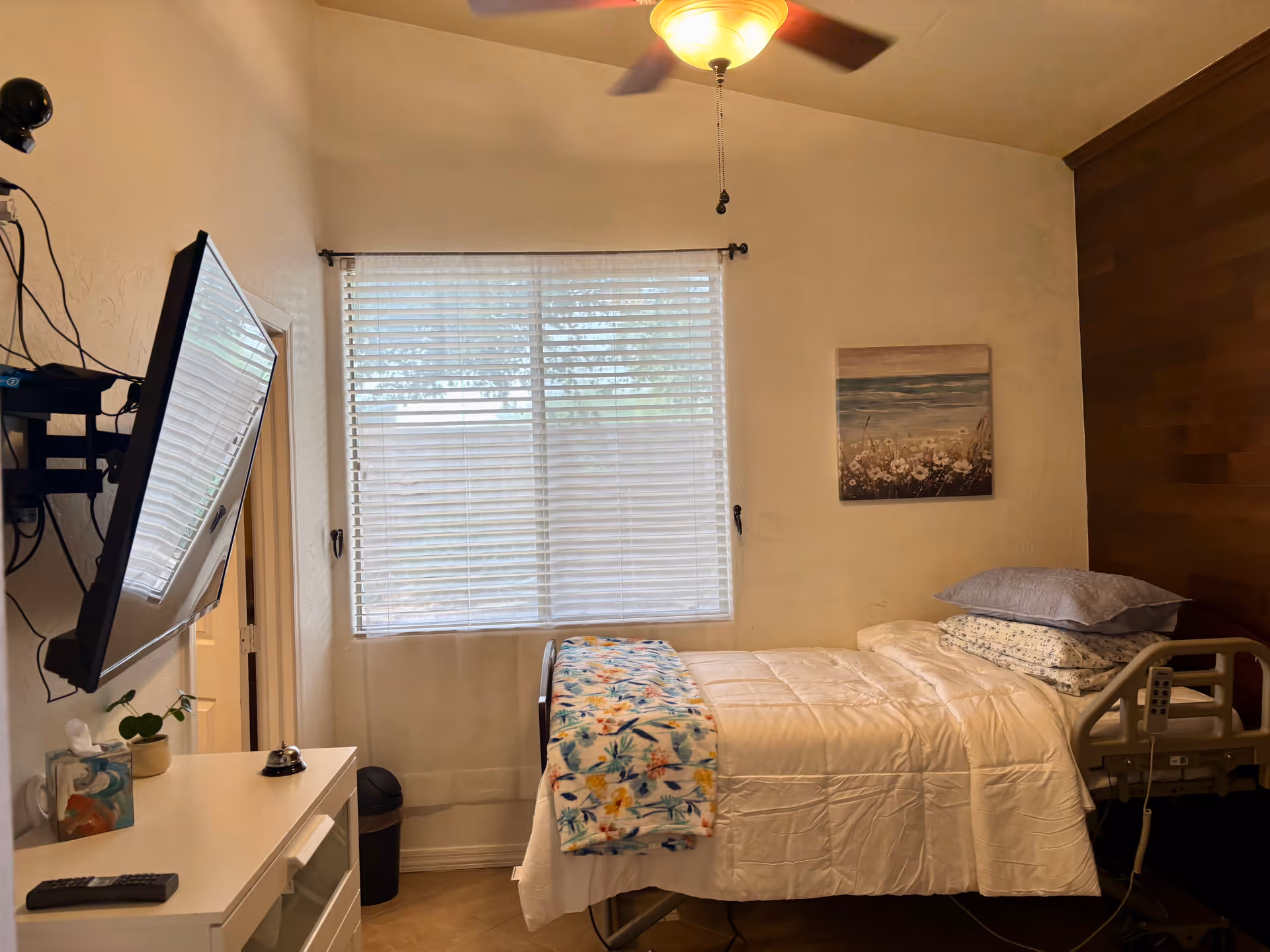 Bedroom in a senior living facility with a bed, mounted TV, blinds, and wall art.