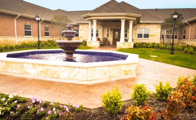 Entrance courtyard of a brick senior living building with a central tiered fountain, landscaped beds, and a covered front porch.