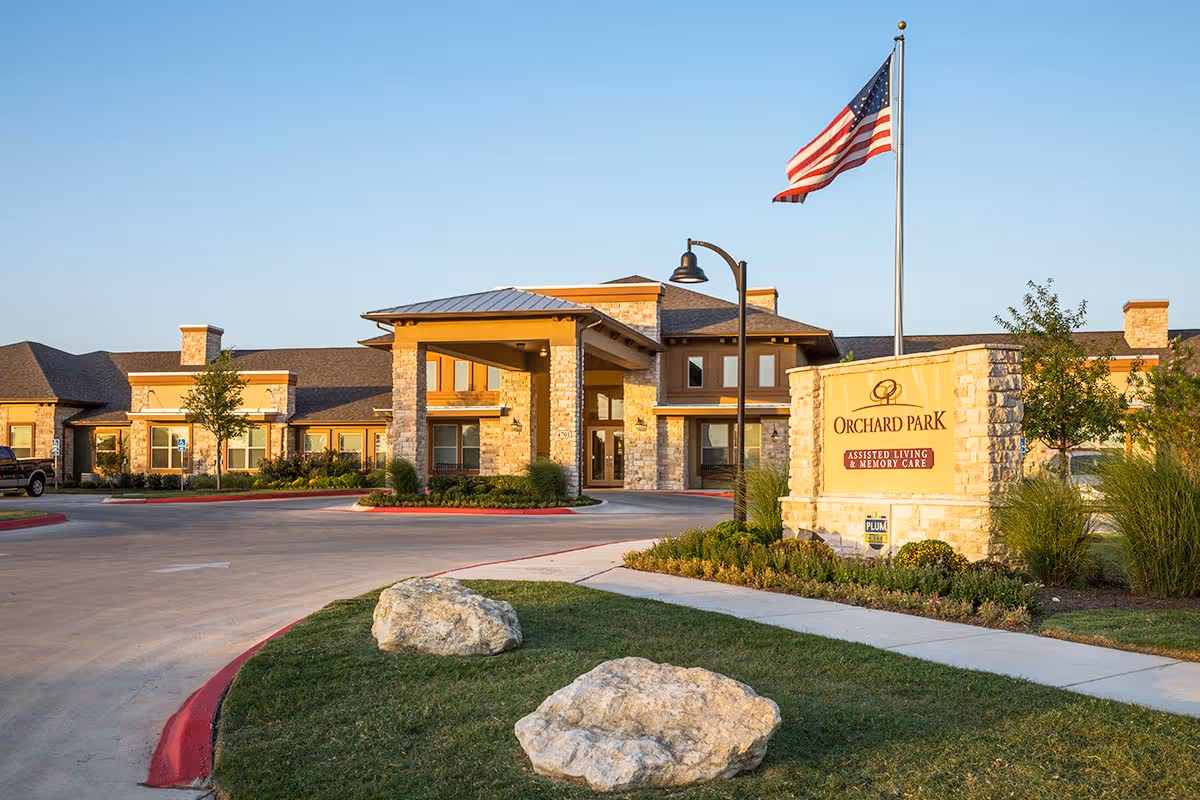 Front entrance of Orchard Park of Kyle assisted living building with a driveway, landscaped grounds, flagpole, and facility sign.