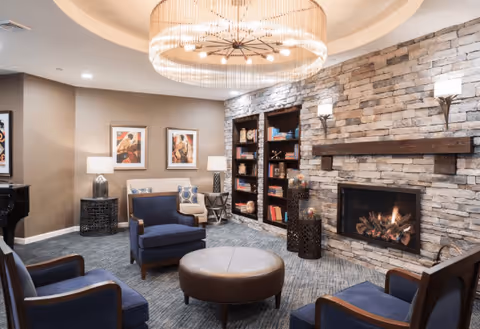 A cozy living room area featuring four blue armchairs arranged around a round brown ottoman. The room has a stone fireplace with a fire burning, built-in bookshelves filled with books and decorative items, two framed artworks on the wall, and a large circular chandelier hanging from a recessed ceiling.