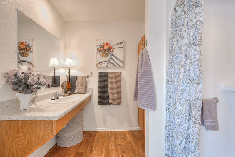 A bright bathroom featuring a long countertop with a sink, a large mirror, and a decorative lamp. There is a vase with flowers on the counter and three towels hanging on the wall. A shower with a patterned curtain is visible on the right side. The floor has a wood-like finish.
