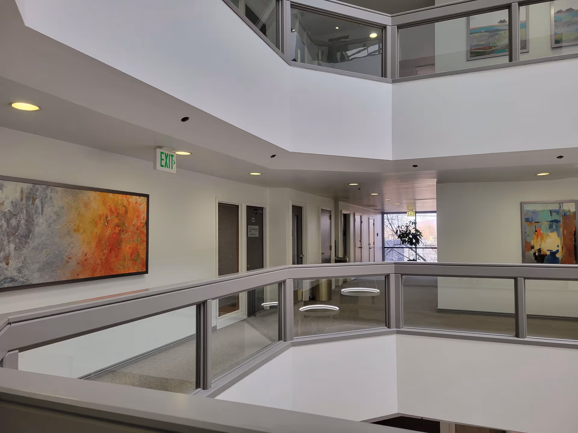 Two-level interior corridor with glass railing, artwork, and an exit sign in a modern facility.