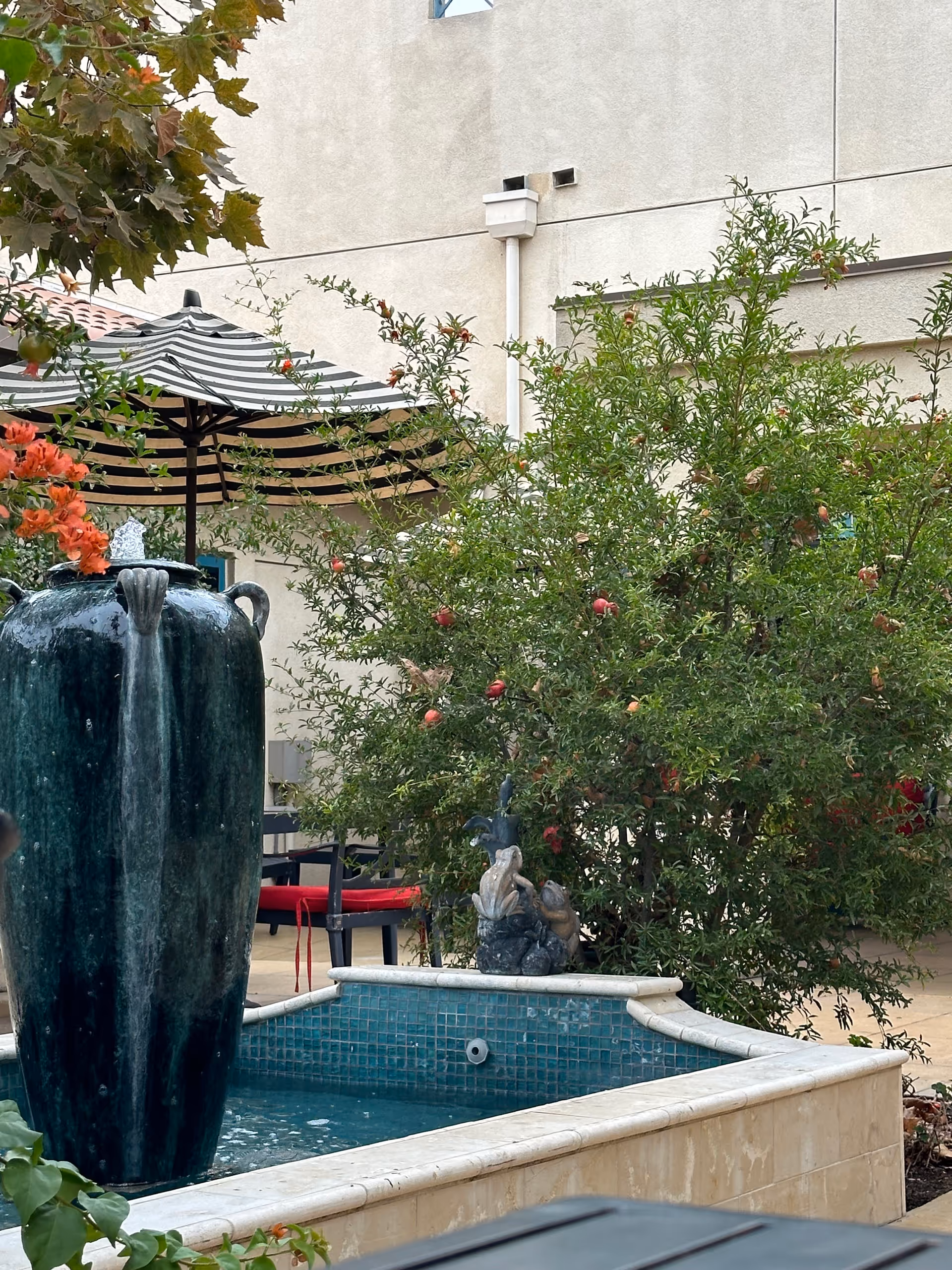 Outdoor courtyard with a tall fountain, umbrella, greenery, and seating near a beige building.