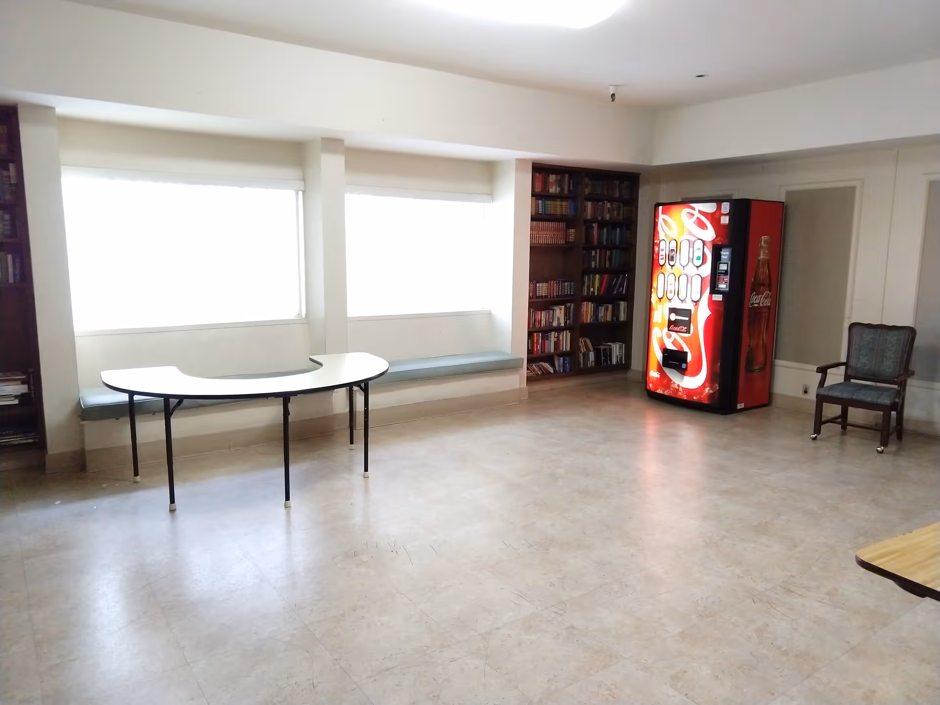 Bright empty common room with a curved table, vending machine, bookshelf, bench, and chair.