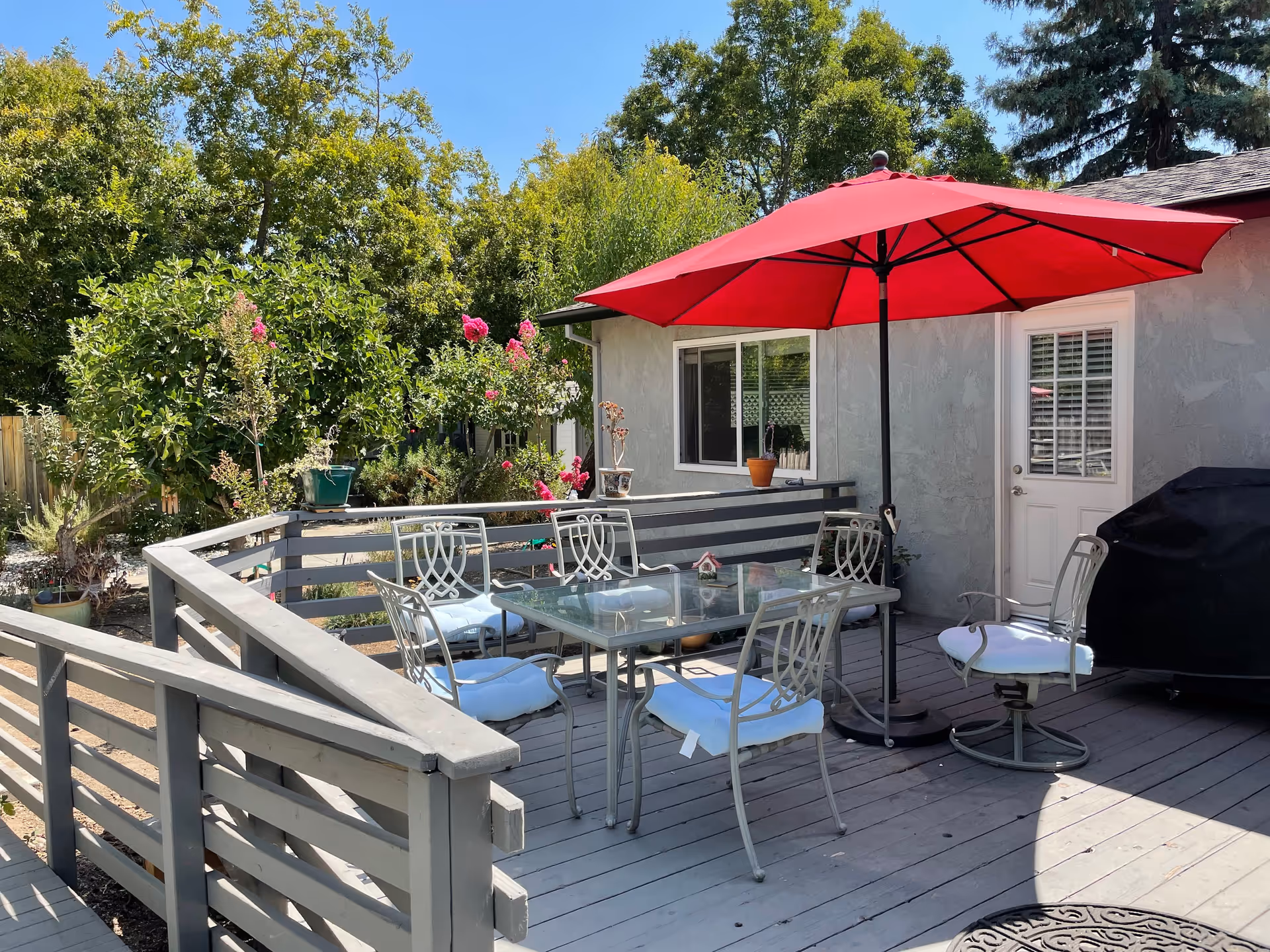 Raised backyard deck with a glass dining table and metal chairs under a red umbrella next to a house door and grill.