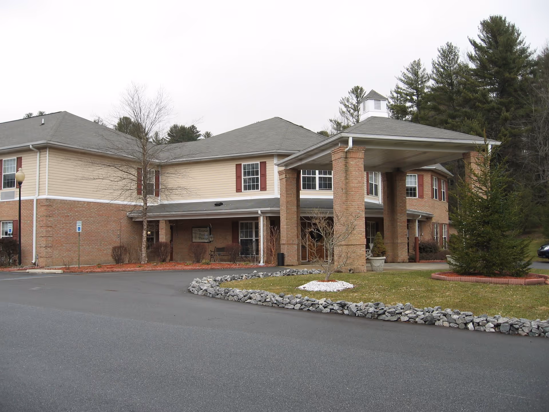 Exterior view of Cedar Mountain House, a two-story building with beige siding and red brick accents. The entrance features a covered driveway supported by brick columns. There is a small landscaped area with rocks and grass in front of the building, and trees are visible in the background.