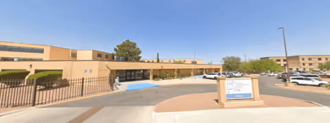 Exterior view of a tan, single-story senior living facility with an entrance sign and a parking lot.