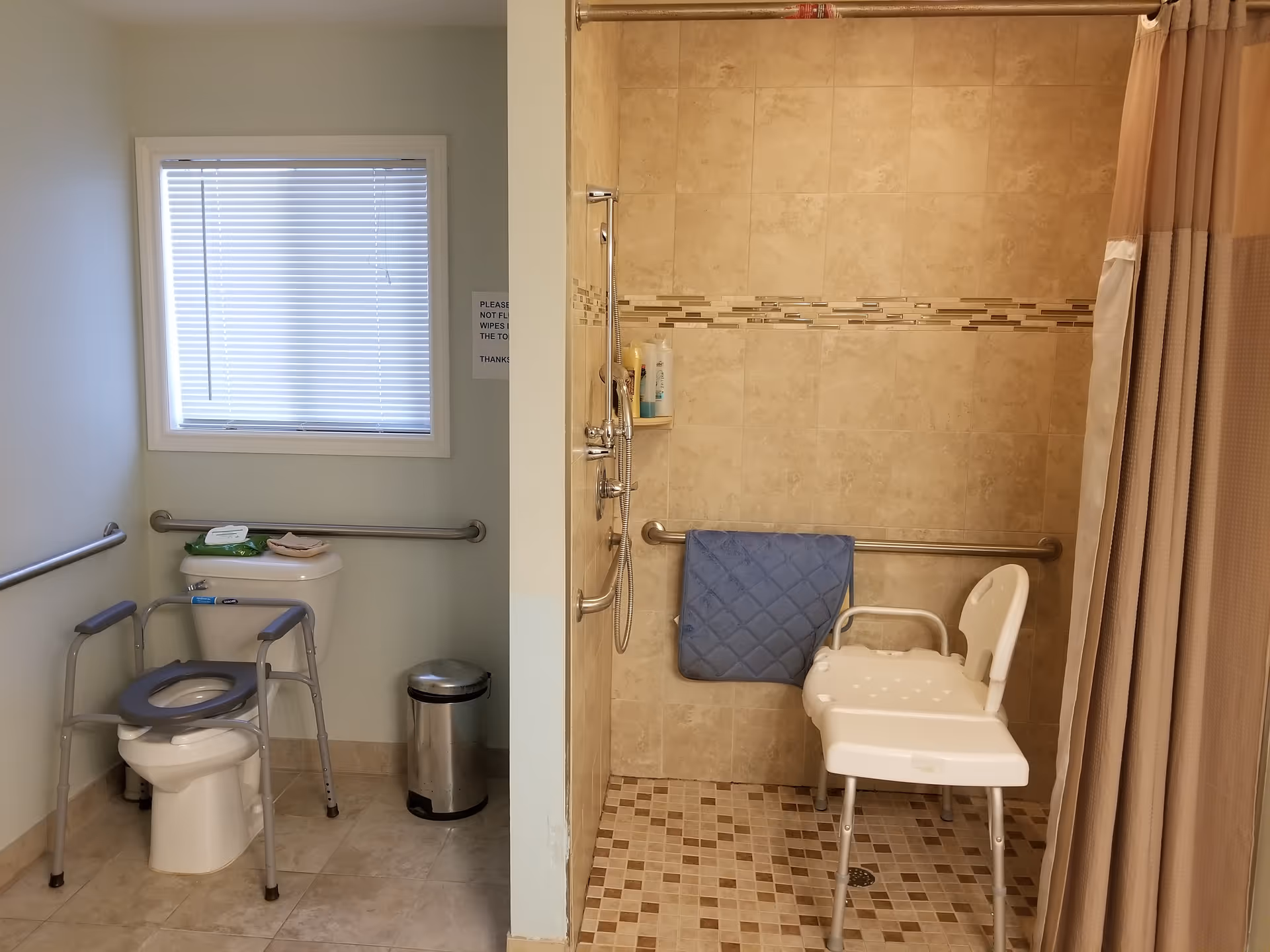 A senior-friendly bathroom with a toilet equipped with a raised seat and support rails, a window with blinds, a small trash can, and a walk-in shower area with beige tiled walls and floor. The shower has a handheld showerhead, a grab bar, a blue towel hanging on the bar, and a white shower chair with armrests. A shower curtain is partially visible on the right side.
