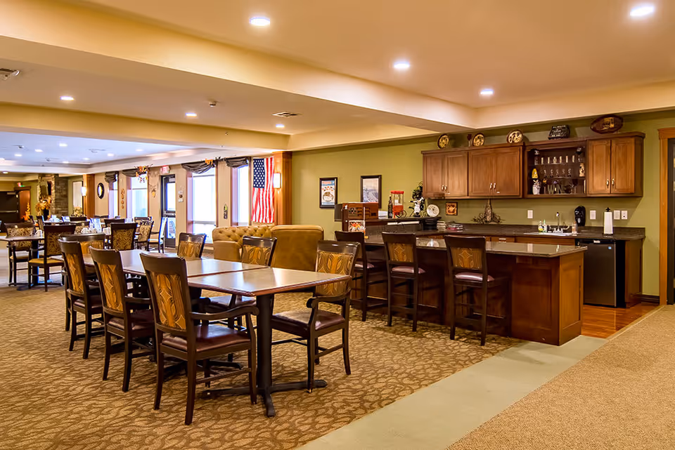 A spacious dining area in a senior living facility with multiple wooden tables and chairs arranged neatly. There is a kitchen counter with bar stools on the right side, wooden cabinets above, and various kitchen items on the counter. The room is well-lit with recessed ceiling lights and has a carpeted floor. An American flag is visible near the windows in the background.