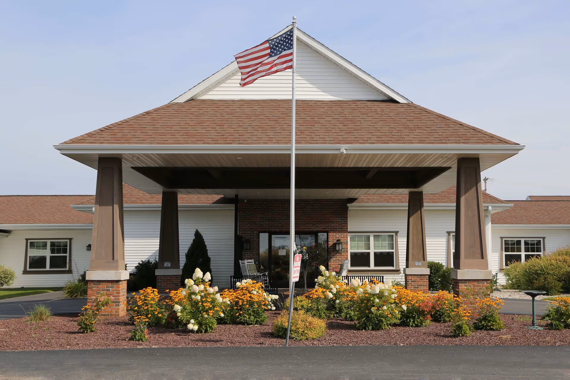 Front entrance of a single-story senior living facility with a covered portico, an American flag on a flagpole, and flowerbeds.