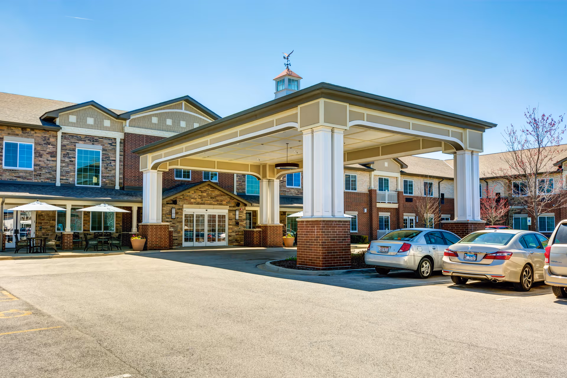Exterior view of Burr Ridge Senior Living facility showing the main entrance with a large covered drop-off area supported by white columns with brick bases. Several cars are parked in front, and there are outdoor seating areas with tables and umbrellas to the left. The building has a mix of brick and stone facade with multiple windows and a clear blue sky above.