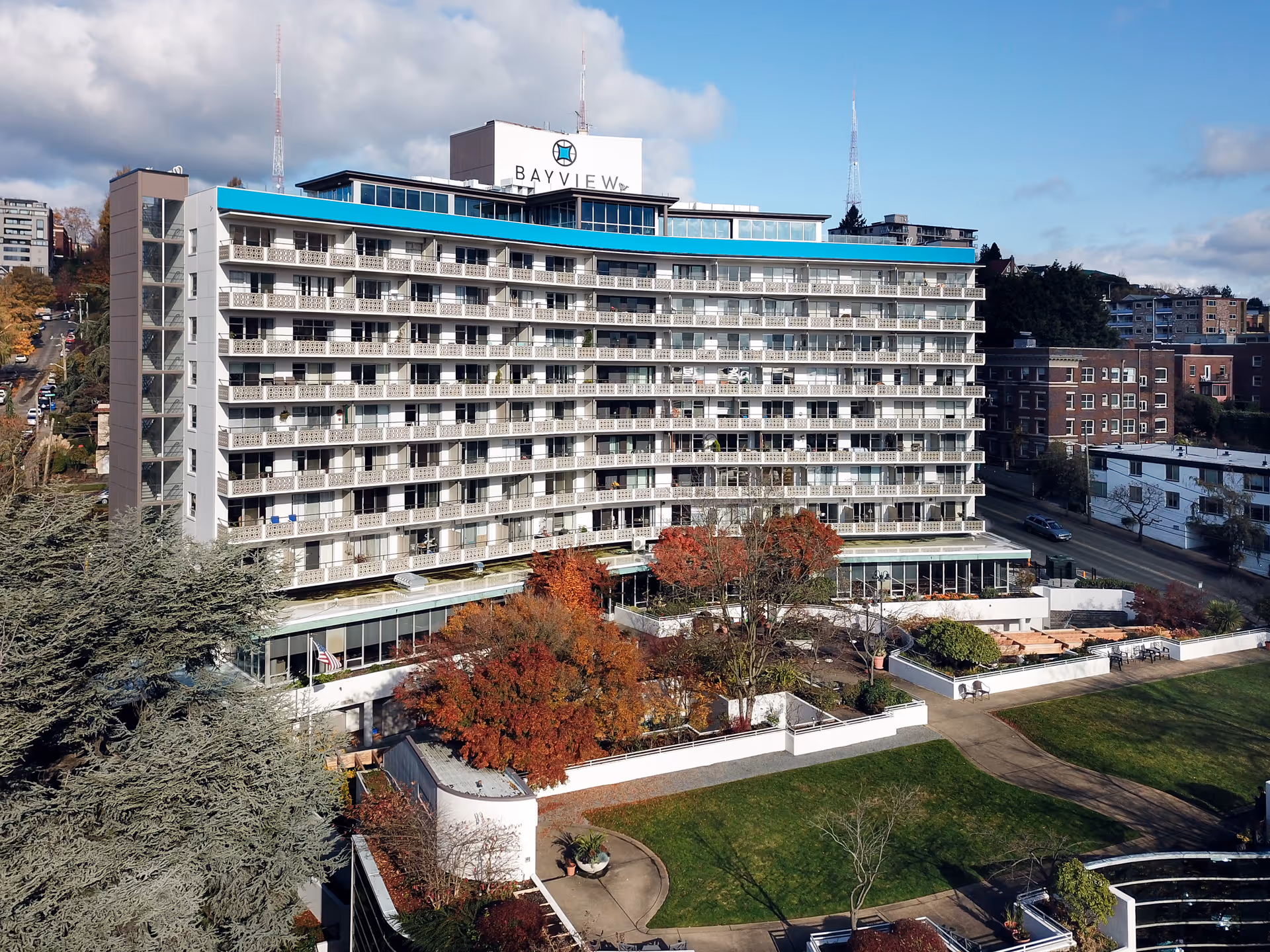 Exterior view of Bayview Retirement Community, a multi-story building with balconies on each floor, surrounded by trees with autumn foliage and a well-maintained lawn in the foreground under a partly cloudy sky.