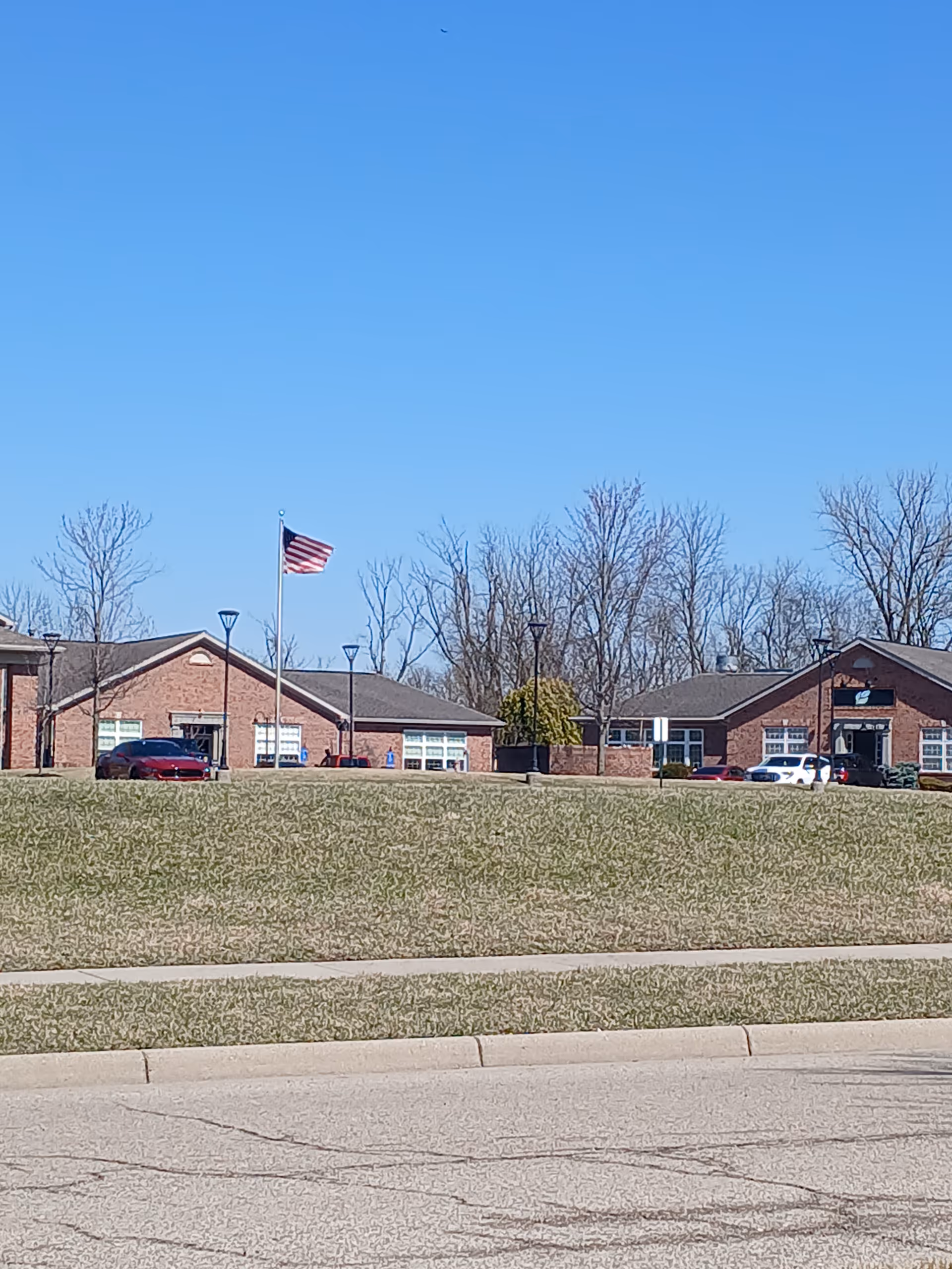Brick hospice building with flagpole, parked cars, leafless trees, and blue sky.