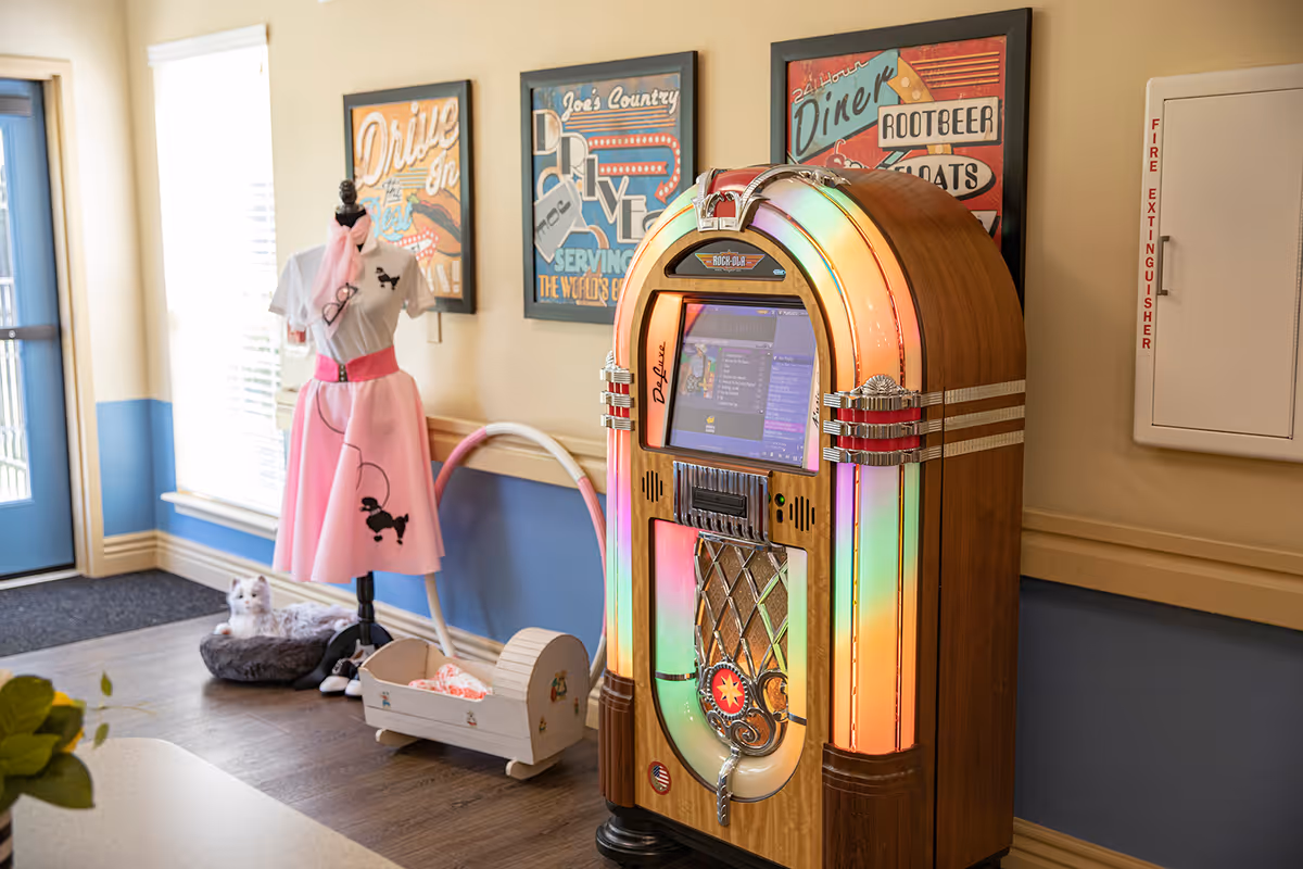 Interior corner of a room featuring a vintage-style jukebox with colorful lights, a mannequin dressed in a 1950s-style pink poodle skirt outfit, a small white cradle, a cat bed with a white cat, and three retro diner-themed framed posters on the wall. A fire extinguisher cabinet is mounted on the wall to the right.