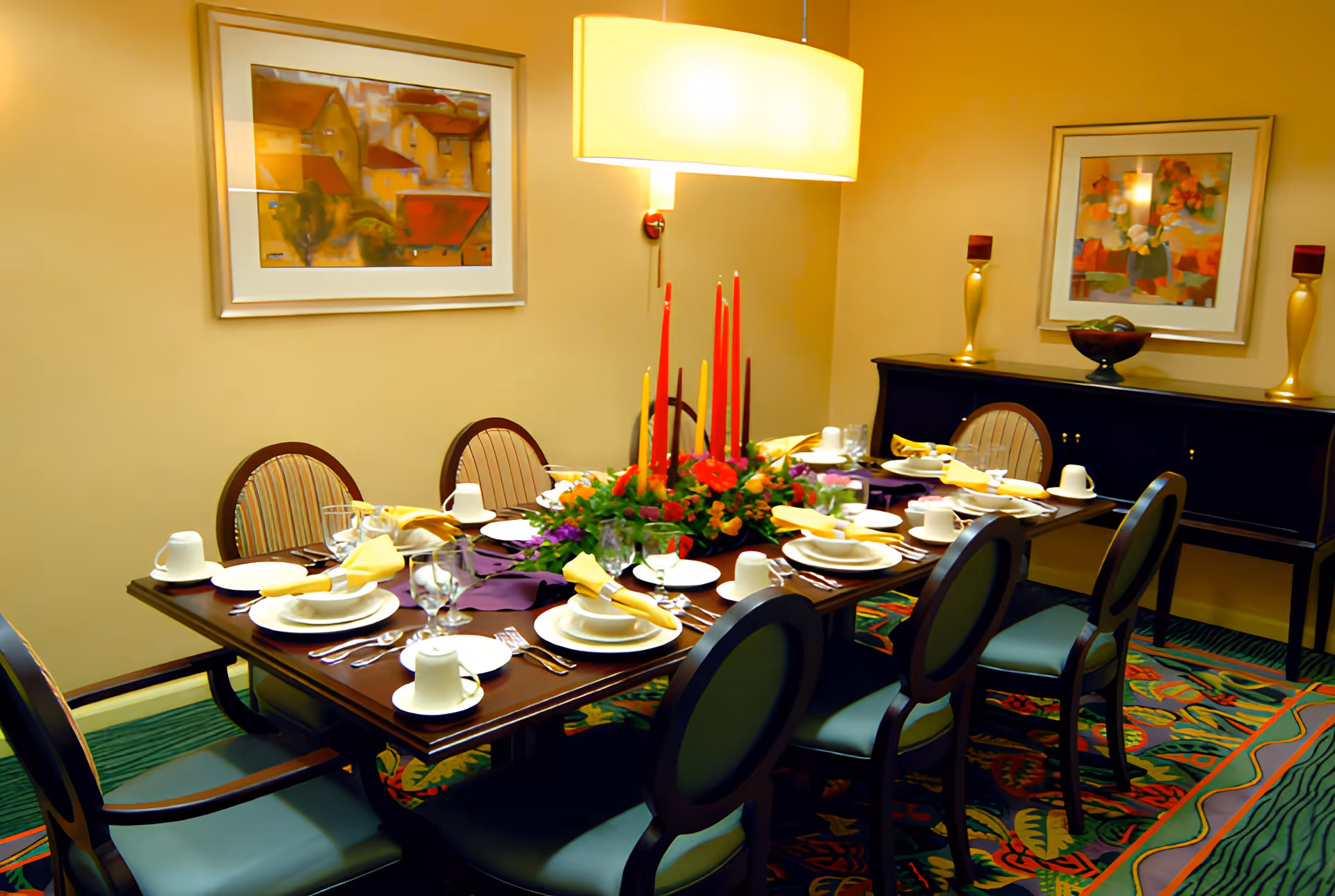 A formal dining room with a long wooden table set for eight people. The table is decorated with a floral centerpiece and tall red and yellow candles. Each place setting includes white plates, cups, glasses, and yellow napkins. The room has beige walls adorned with two framed abstract paintings, a dark wooden sideboard with decorative items, and a colorful patterned carpet. A large rectangular pendant light hangs above the table.