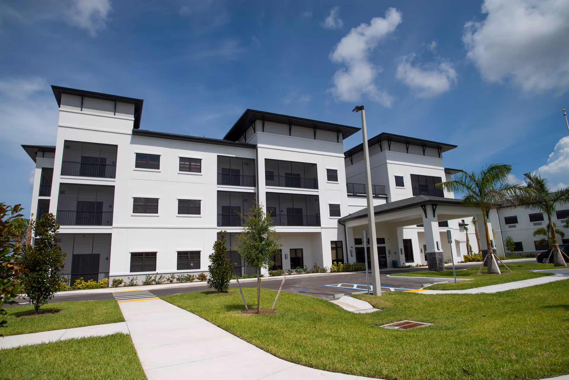 Exterior view of a modern three-story senior living facility building with white walls and black window frames, surrounded by green grass, palm trees, and a clear blue sky with some clouds.