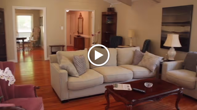 A cozy living room with a beige sofa adorned with multiple cushions, a wooden coffee table with a remote control and a newspaper on it, a side table with a lamp, and a painting on the wall. The room has wooden flooring and opens into other rooms with visible furniture.