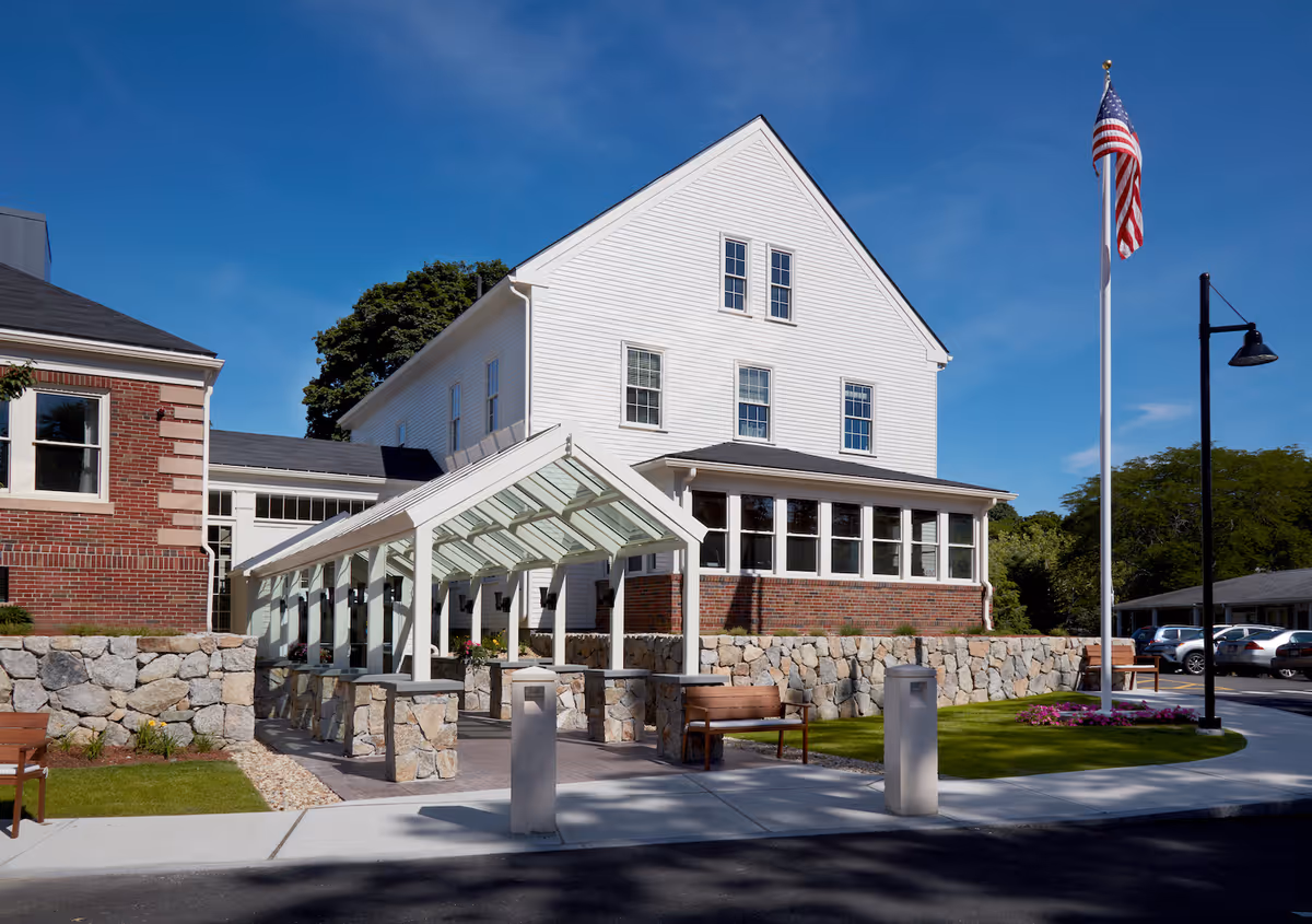 Front exterior of a multi-story white and brick senior living building with a covered glass walkway, stone walls, benches, and an American flag.