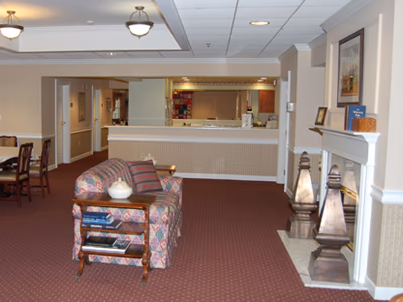 Carpeted senior living common room with patterned sofas, a fireplace mantel, and a reception desk in the background.