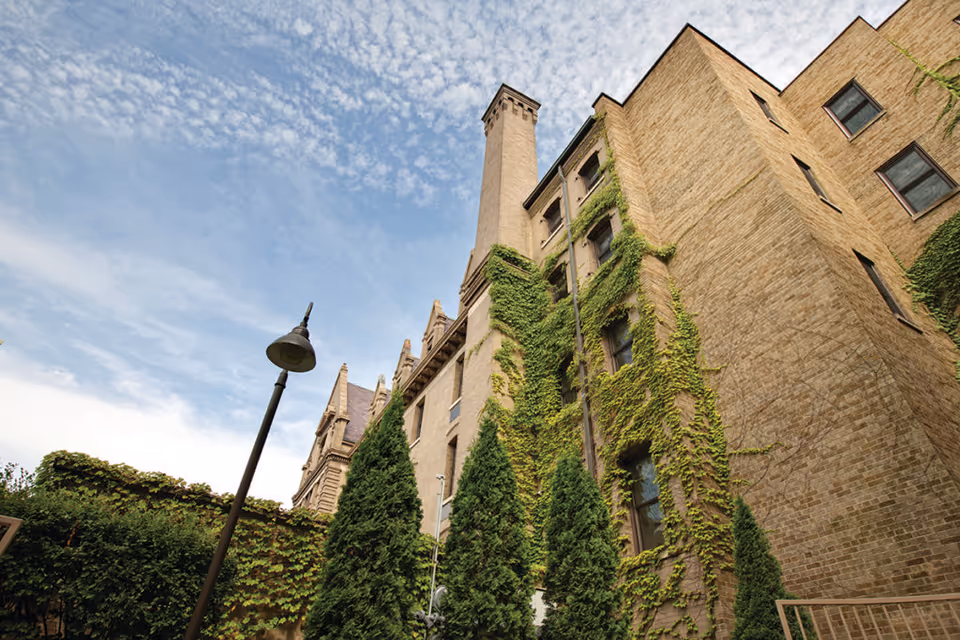 Ivy-covered brick building exterior with a tall chimney, evergreen trees, and a lamppost under a partly cloudy sky.