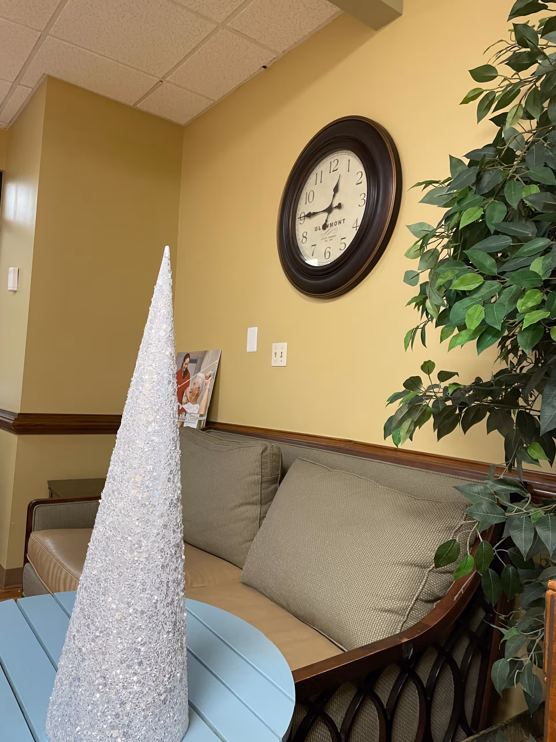 Senior living lounge area with beige walls, clock, plant, and cushioned couch.