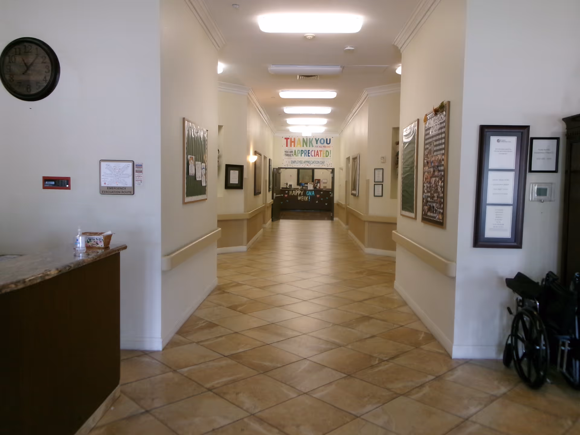 Interior hallway of a healthcare facility with tiled floors and beige walls. The hallway is well-lit with ceiling lights and has handrails on both sides. On the left side, there is a reception desk with a clock above it and a hand sanitizer bottle. On the right side, there are framed documents and a wheelchair. At the end of the hallway, a colorful banner reads 'THANK YOU APPRECIATED!' and 'HAPPY CNA WEEK!'.