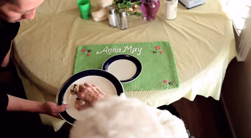 An elderly person reaching for a snack on a plate held by another person at a table with a green placemat embroidered with the name 'Anna May'. The table is covered with a yellow tablecloth and has a small flower vase and salt and pepper shakers.