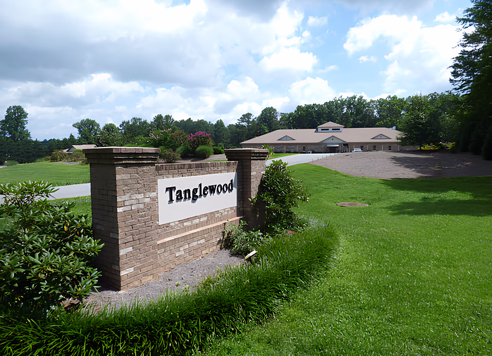 Landscaped entrance with a brick sign reading "Tanglewood" and a single-story facility building in the background under a partly cloudy sky.