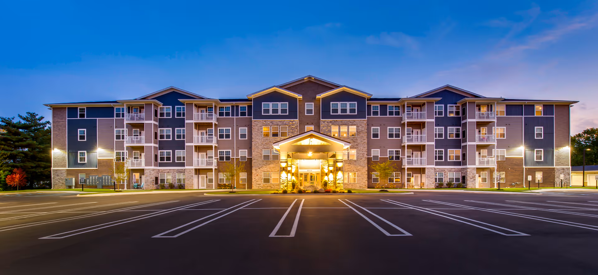 Four-story apartment building with balconies and a lit central entrance seen from an empty parking lot at dusk.