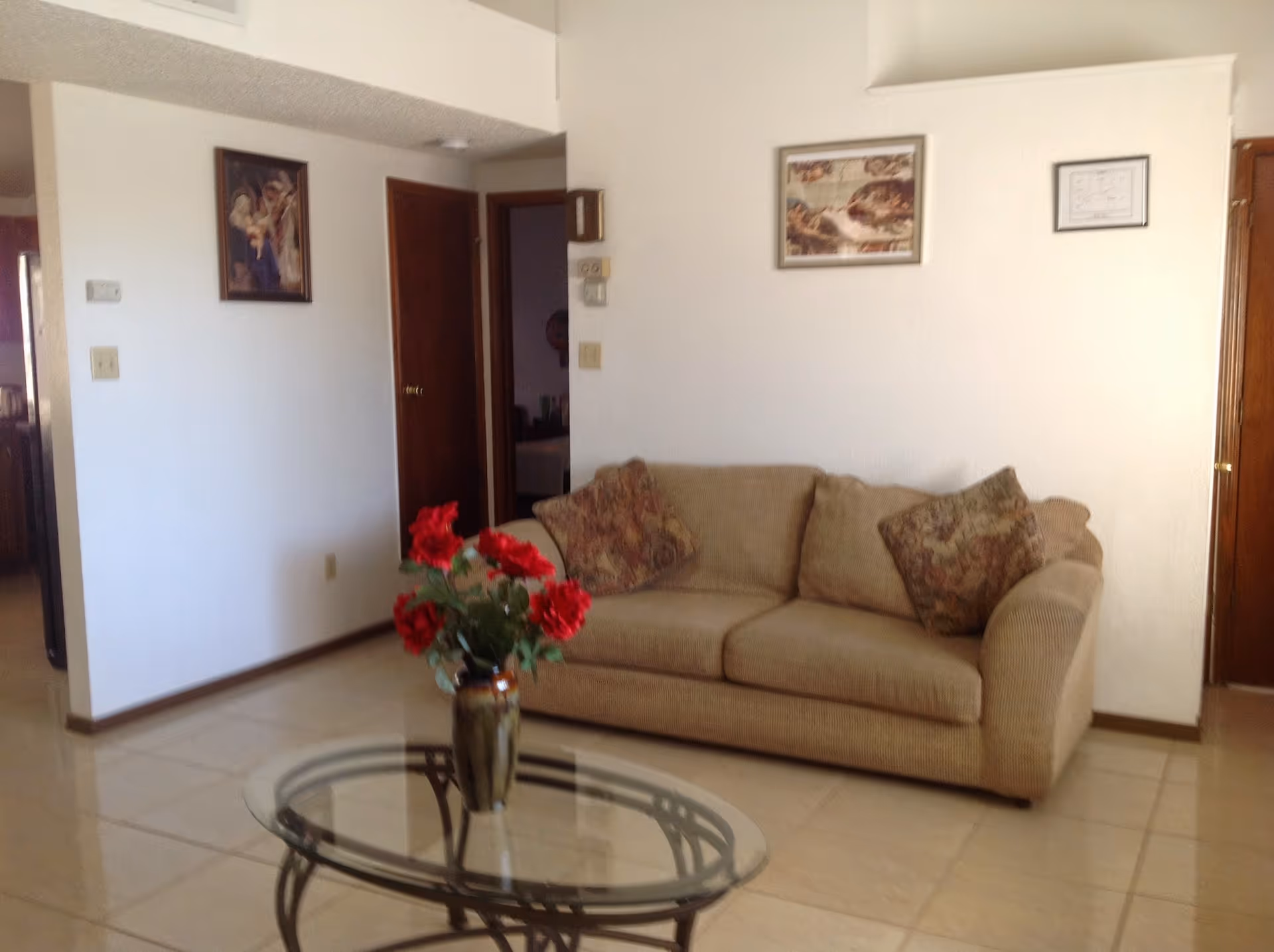 Beige sofa with decorative pillows beside a glass coffee table with a vase of red flowers.