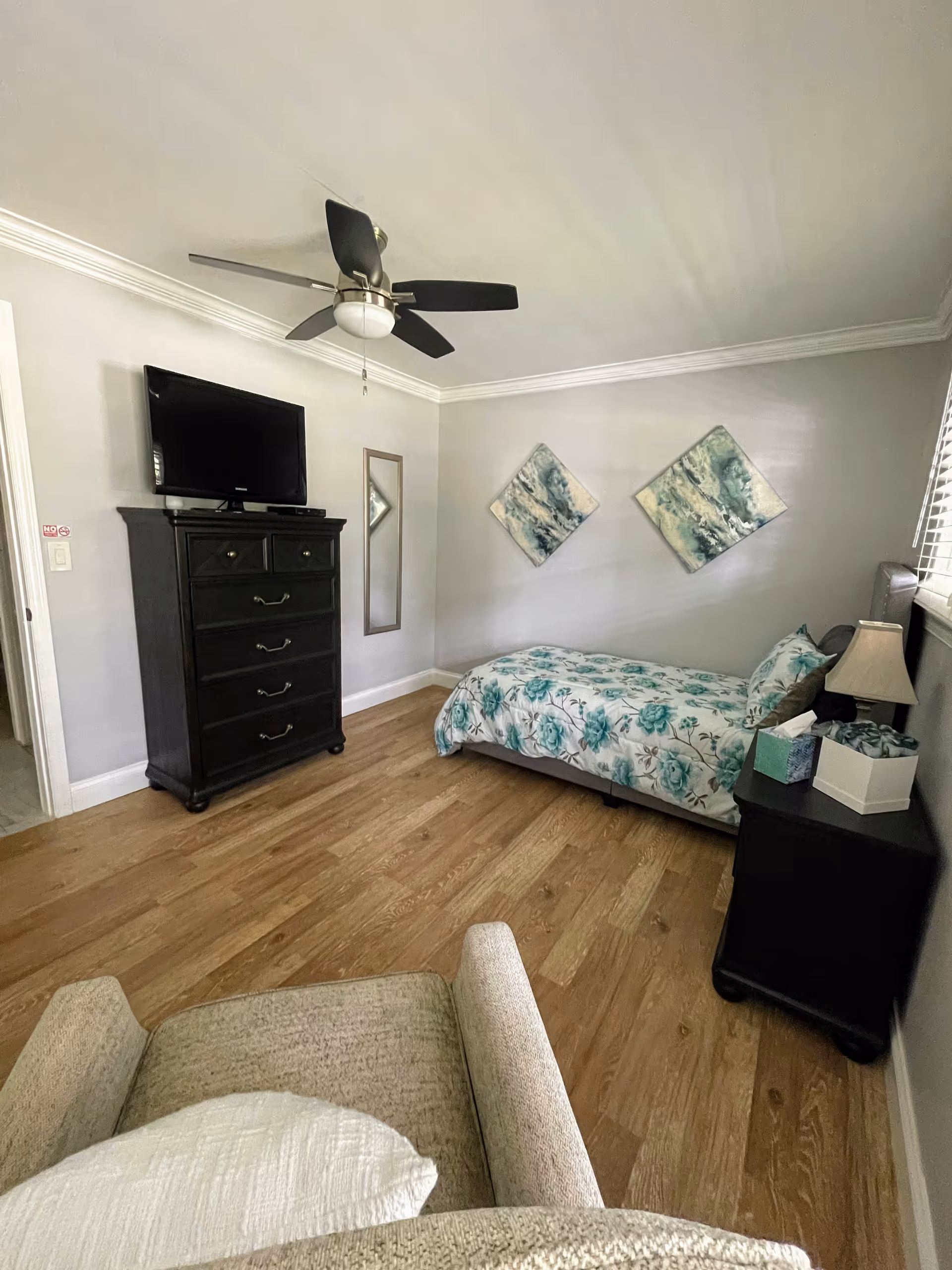 Single bed with floral bedding, dresser with TV, ceiling fan, and wall art.
