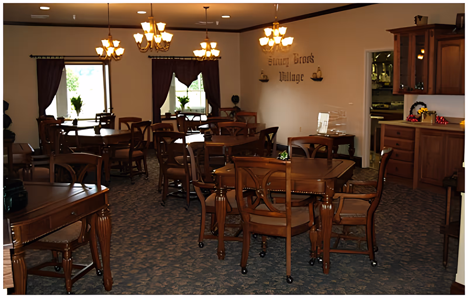 A dining room with multiple wooden tables and chairs arranged neatly. The room has carpeted flooring, several chandeliers hanging from the ceiling, and windows with dark curtains letting in natural light. There is a wooden cabinet on the right side and a sign on the wall that reads 'Stoney Brook Village'.
