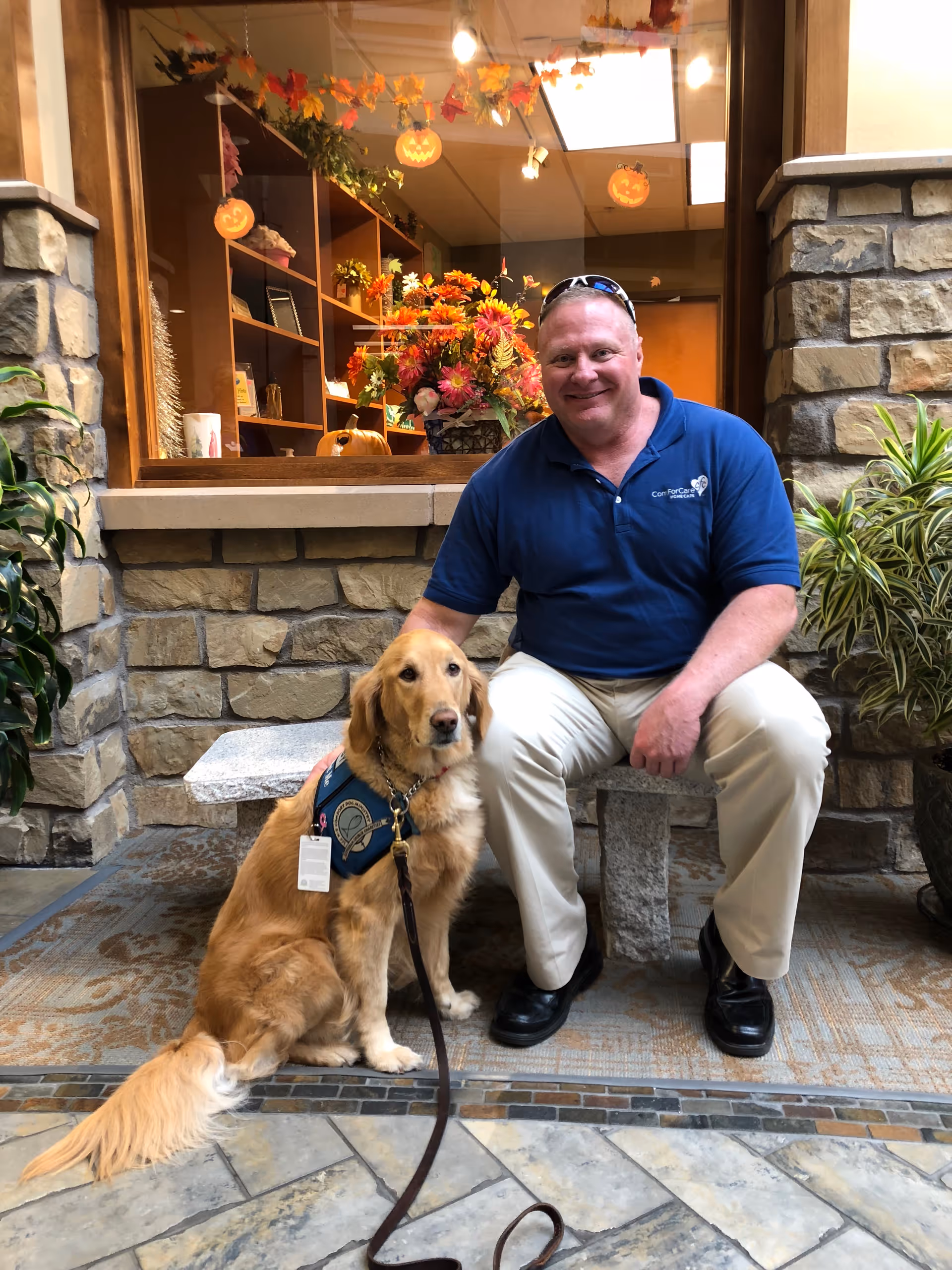 A man in a blue polo sits outside with a golden retriever wearing a vest.