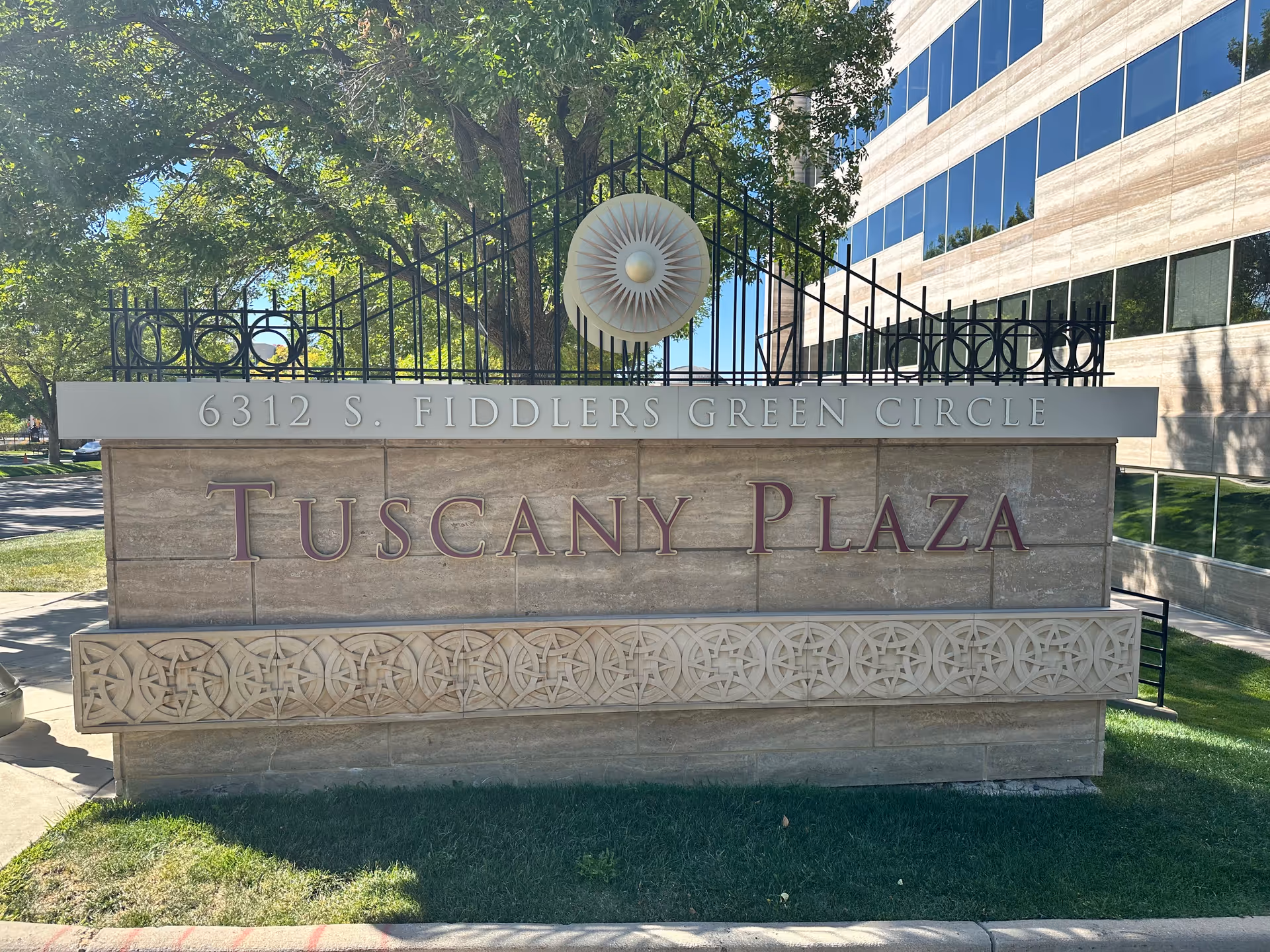Stone entrance sign reads 6312 S. Fiddlers Green Circle for Tuscany Plaza with a decorative fence.
