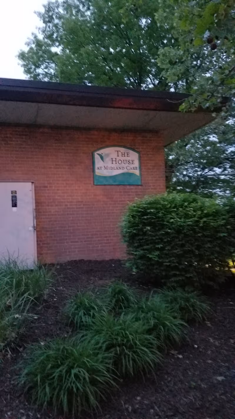 Brick building exterior with sign reading The House at Midland Care, surrounded by shrubs and trees.