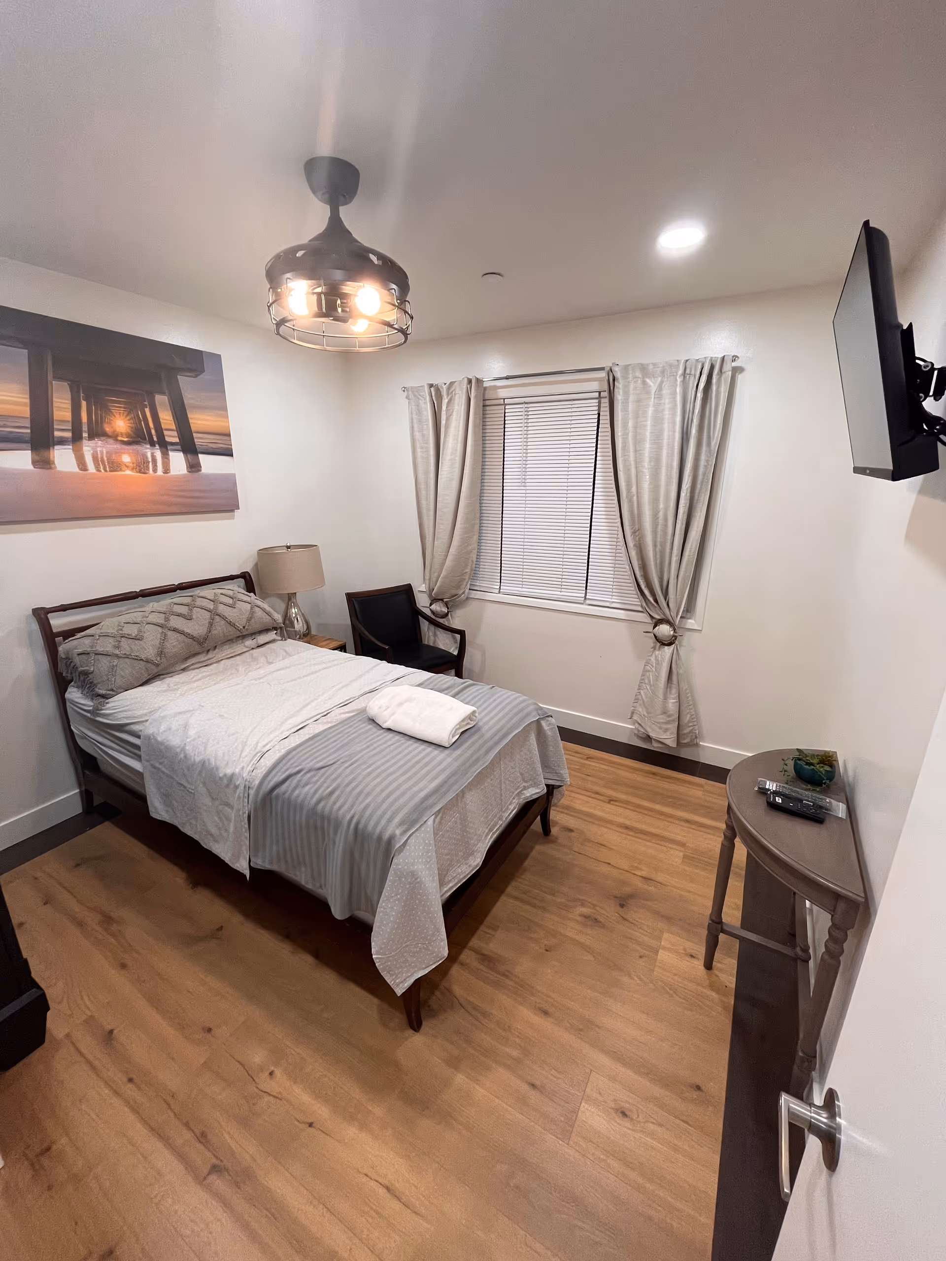 A neatly furnished bedroom with bed, chair, window, and mounted TV.
