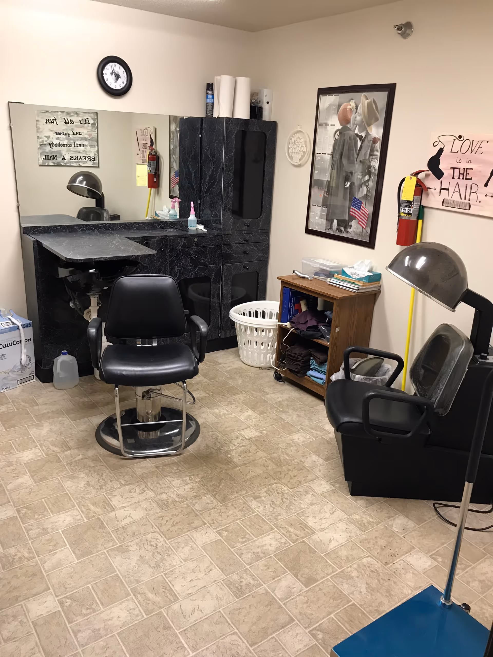 Small hair salon room with a styling chair, hooded dryer, mirror, cabinets, and wall decor.