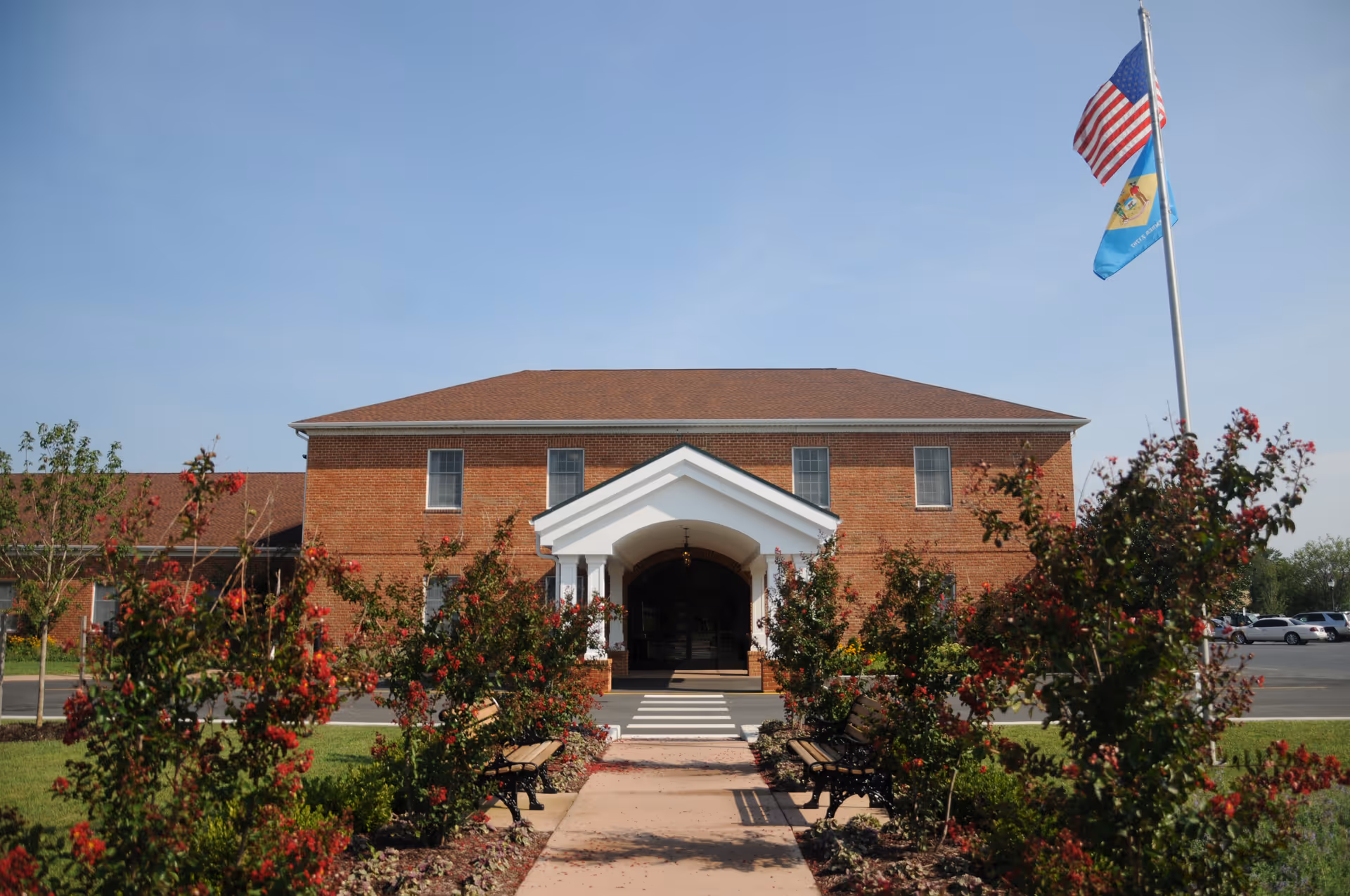 Front entrance of a two-story brick building with a white portico, walkway, benches, landscaped shrubs and flags.