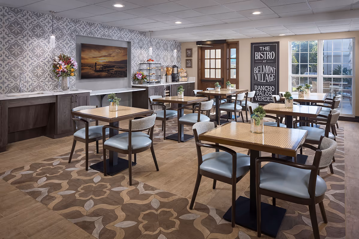 Empty dining room with wooden tables, matching chairs, buffet area, and Belmont Village Bistro sign.
