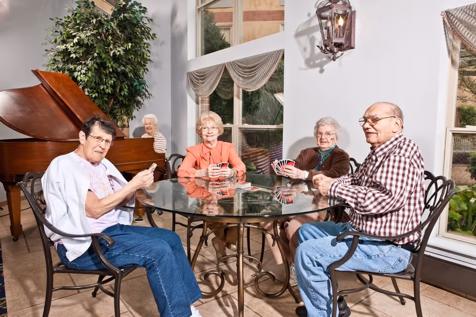 Five elderly individuals sitting around a glass table playing cards in a bright room with large windows, a grand piano, and a potted plant in the background.