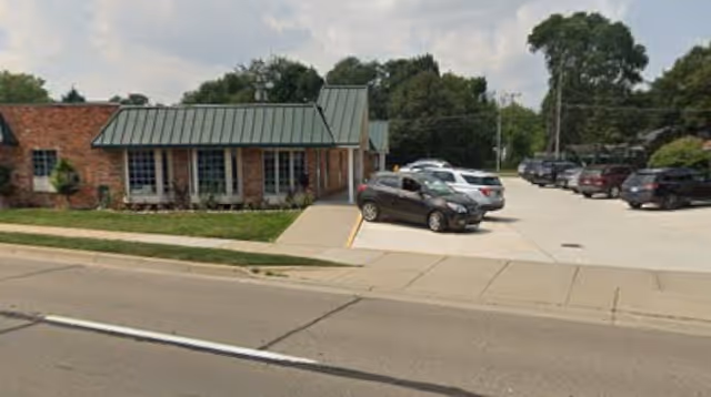 Front view of a senior care facility with brick walls, green roof, and parking.