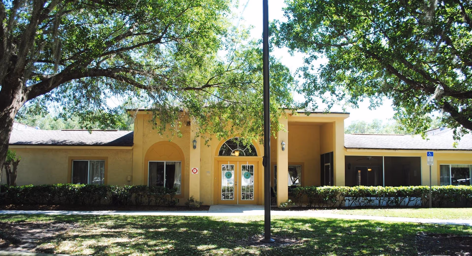 Single-story yellow building entrance with double glass doors, shrubs, large shade trees, and a central lamp post in front.
