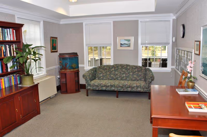 A cozy sitting room in a senior living facility featuring a floral-patterned sofa against a wall with two windows. To the left, there is a wooden bookshelf filled with books and a potted plant on top. Next to the sofa is a fish tank on a wooden stand. On the right side, there is a wooden table with chairs, a few books, and a small potted orchid. The walls are decorated with framed artwork, and a clock is mounted on the wall above the table.