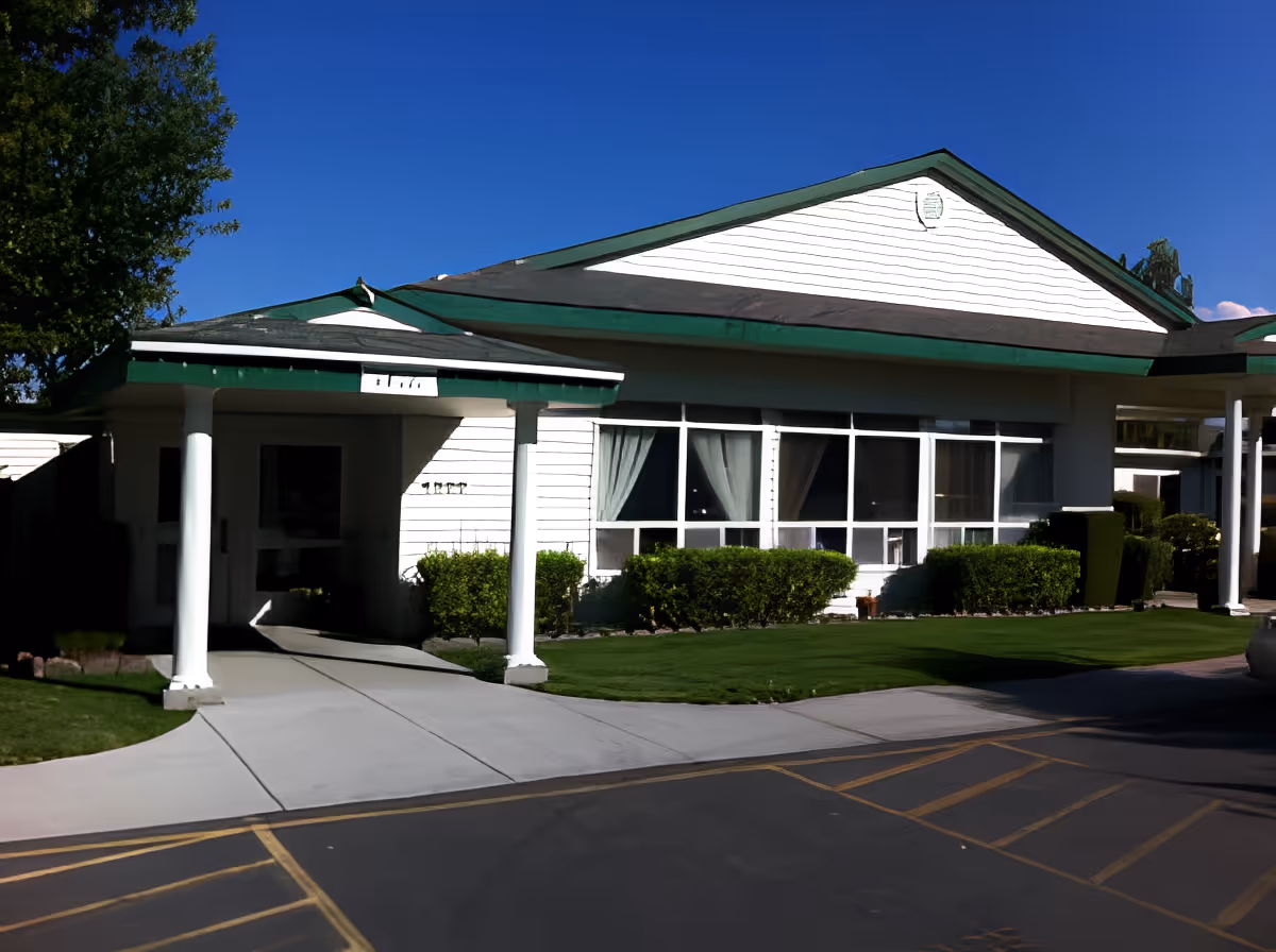 Exterior view of Meadowbrook Place Assisted Living building with white walls, green roof trim, large windows with curtains, a covered entrance supported by white columns, and neatly trimmed bushes and lawn under a clear blue sky.