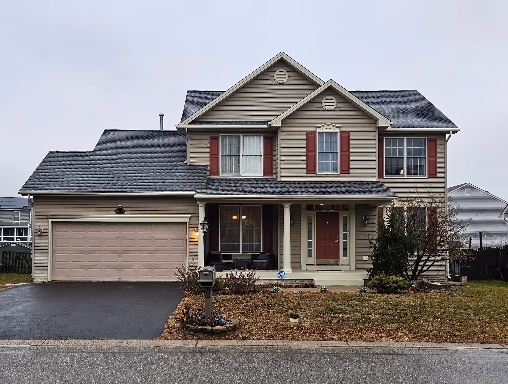 Two-story beige house with a two-car garage, red shutters, and a front yard.